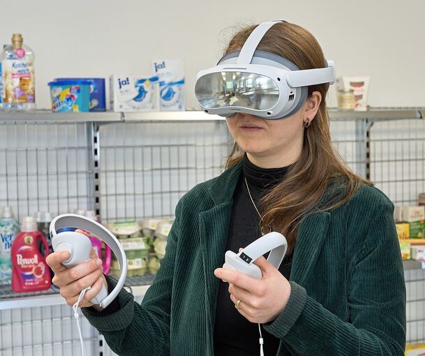 Maike Burkhardt, Studienrätin an der Erich-Bracher-Schule, in einem Unterrichtsraum mit VR-Brille auf dem Kopf und Supermarktregalen im Rücken. Fotos: Andreas Becker