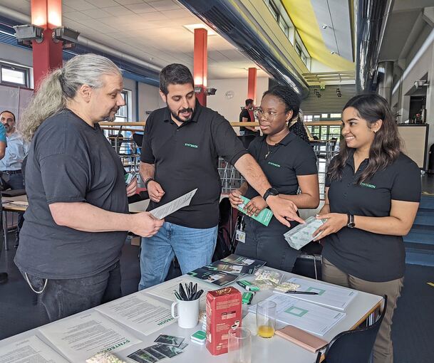 Jörg Wiesner (links) informiert sich bei der Jobmesse in der BorgWarner-Kantine am Stand des Unternehmens Syntegon. Ioannis Michailidis, Anastasia Ansong und Sharika Chandrasegaran beraten den BorgWarner-Mitarbeiter zu Stellen bei dem Sondermaschinen