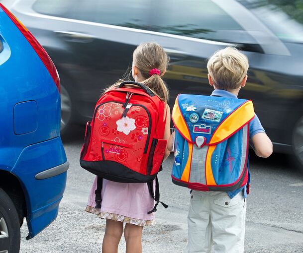 Zwei Kinder mit Schulranzen auf dem Rücken schauen ein vorbeifahrendes Auto an.