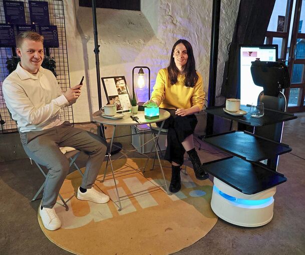 Julian Kemmer und Lena Ahner sitzen in der Alten Kelter in Bietigheim-Bissingen in der Gastronomie-Ecke der Ausstellung. Rechts steht ein Service-Roboter; der Würfel auf dem Tisch leuchtet dann grün, wenn der Gast auf ihn getippt hat; der Ober erhält