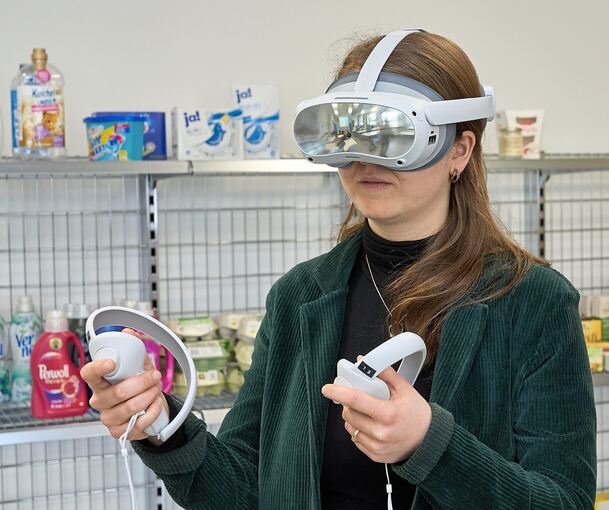 Maike Dasch, Studienrätin an der Erich-Bracher-Schule und deren Pressebeauftragte, in einem Unterrichtsraum mit VR-Brille auf dem Kopf und Supermarktregalen im Rücken.