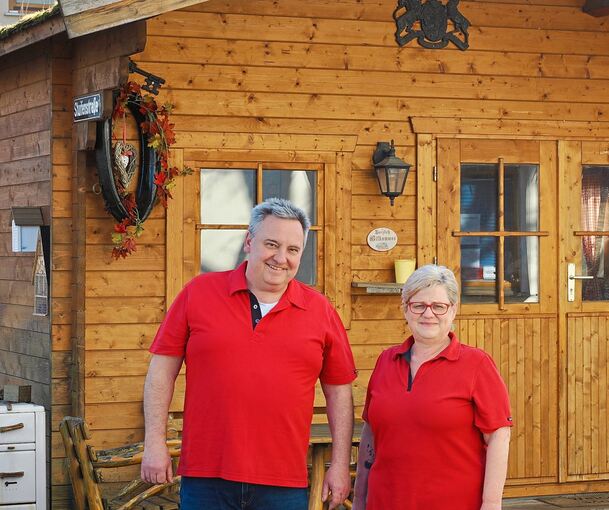 Ralf und Andrea Holzwarth vor der Holzhütte neben ihrem Wohn- und Geschäftsgebäude. Dort kann die Kundschaft weiterhin verschiedene Speisen aus dem Imbiss zu sich nehmen.
