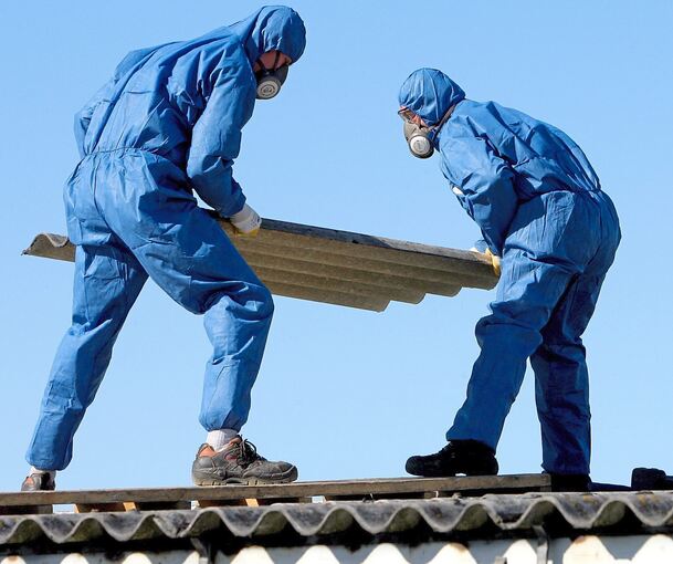 Nur etwas für Profis: Arbeiter in Schutzanzügen und mit Luftfiltern über Mund und Nase entfernen Asbestplatten vom Dach eines alten Gebäudes.