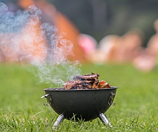 Ein rauchender Grill mit Fleisch steht mittig auf dem grünen Rasen.