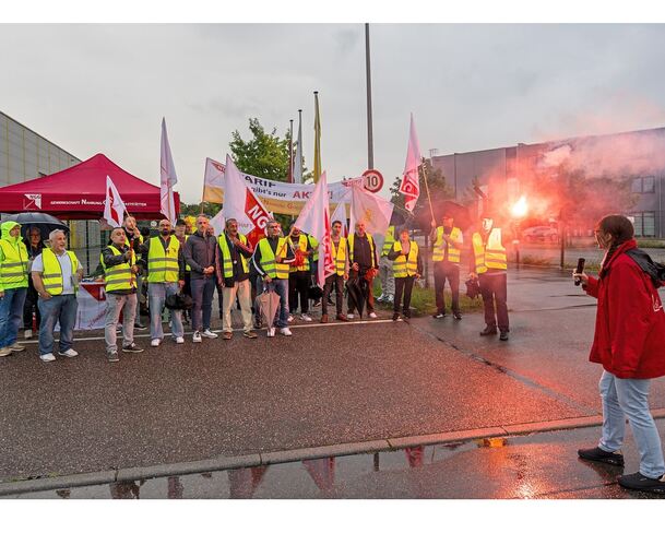 Warnstreik in gelben Westen und mit Bengalo: Lieken-Mitarbeiter der Frühschicht stehen am Montagmorgen in einer kurzen Regenpause vor dem Werksgelände. Rechts (mit Mikrofon) Magdalena Krüger, Geschäftsführerin der NGG-Region Stuttgart, links der Bild