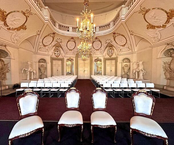 Die Wände sind mit Ornamenten geschmückt. Im Bildvordergrund stehen vier mit Samt bezogene Stühle. Weiter hinten im Saal befinden sich mehrere Stuhlreihen.