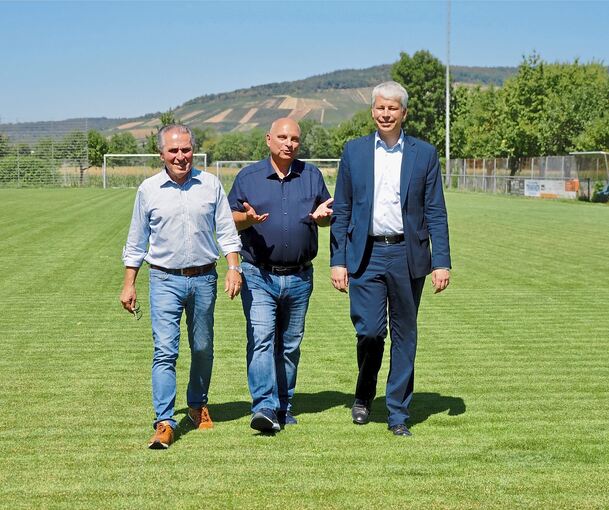 Bundesverkehrsminister Steffen Bilger (von rechts), Bürgermeister Jürgen Scholz und Umweltexperte Claus-Peter Hutter spazieren über den grünen, mit Mineralwasser gegossenen Sportplatz. Im Hintergrund sind vertrocknete und brachliegende Weinbergfläche