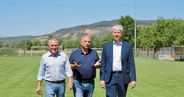 Bundesverkehrsminister Steffen Bilger (von rechts), Bürgermeister Jürgen Scholz und Umweltexperte Claus-Peter Hutter spazieren über den grünen, mit Mineralwasser gegossenen Sportplatz. Im Hintergrund sind vertrocknete und brachliegende Weinbergfläche